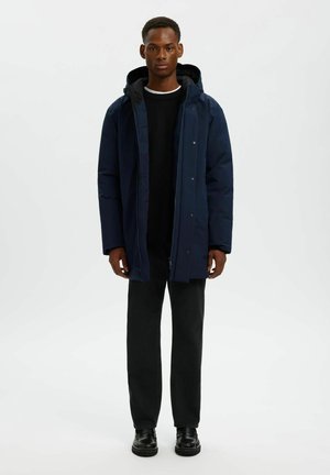 Parka bleu marine avec une capuche, disposant d'une fermeture éclair et de boutons-pression, de poches latérales et d'un tissu lisse et légèrement isolé.