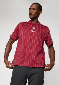 Mand iført en maroon stribet Adidas t-shirt med hvide striber på ærmerne og sorte bukser mod en ensfarvet grå baggrund.