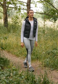 Juodas pūkuotas liemenė virš pilkos ilgomis rankovėmis palaidinės, derinama su šviesiai pilkomis leggingais. Apranga apima pilkus sportinius batus ir yra nustatyta natūralioje aplinkoje.