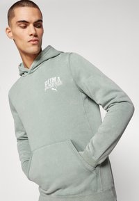 Green hoodie made of soft fabric, features a front pouch pocket, and white printed "PUMA sports club" logo on the chest.