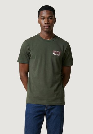 Giovane uomo in piedi, che indossa una t-shirt Colmar verde oliva con logo e jeans blu, su uno sfondo chiaro e semplice.