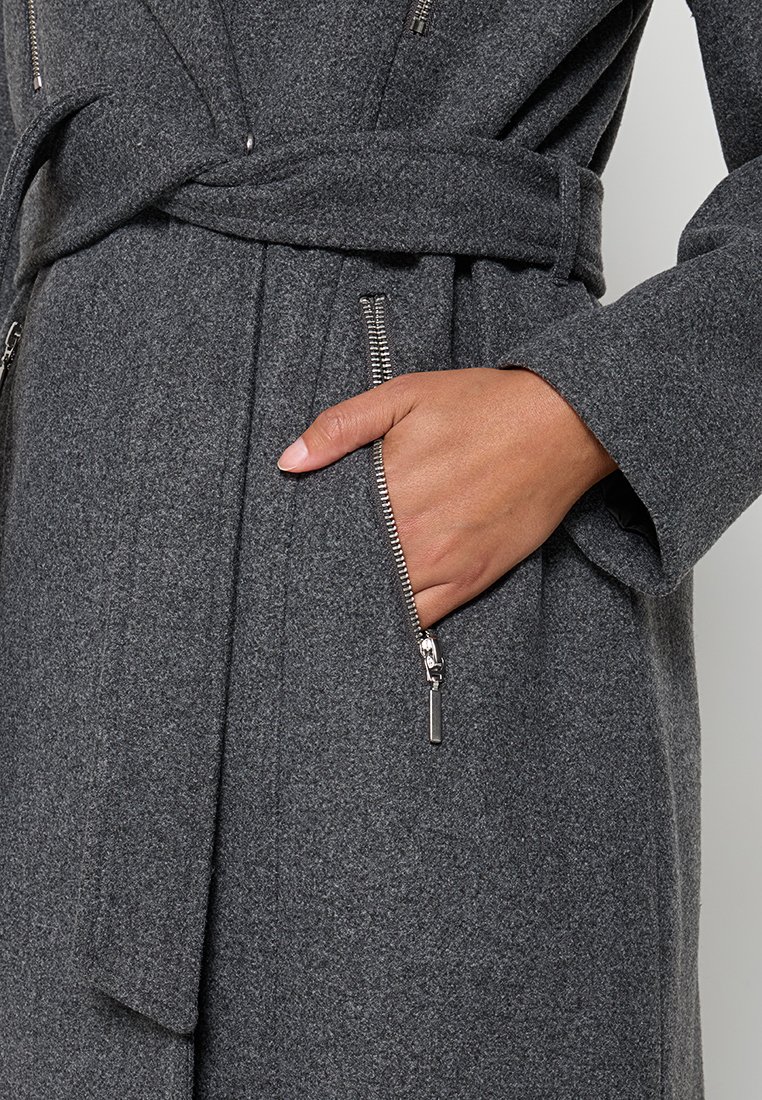 Main dans la poche d'un manteau en laine gris foncé avec une fermeture éclair argentée visible et un détail de ceinture à la taille.