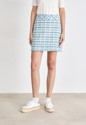 Femme portant une mini-jupe bleue et blanche à motifs, un top blanc à manches courtes, des chaussettes blanches et des baskets blanches, debout sur un sol uni.