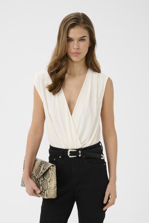 Kvinde med langt brunt hår iført en ærmeløs hvid slå-om top, sorte bukser med bælte, holder en beige clutch med slangeskind.