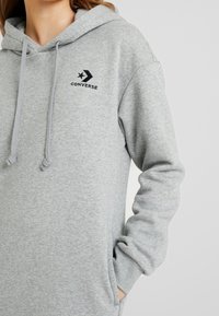 Sudadera gris hecha de un tejido suave y texturizado. Presenta un bolsillo tipo canguro, cordones y un logo negro con una estrella en la parte frontal.