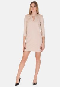 Vestito beige con maniche a tre quarti, scollo a goccia con apertura e design aderente, abbinato a décolleté nere. Tessuto dalla texture liscia.