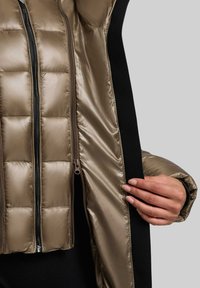 Veste rembourrée avec un extérieur brillant de couleur bronze, présentant un motif matelassé, des manches noires et une fermeture éclair à l'avant.
