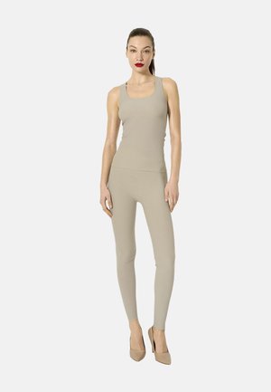 Donna in piedi con indosso un top beige senza maniche, pantaloni aderenti abbinati e scarpe beige con tacco alto su sfondo chiaro uniforme.
