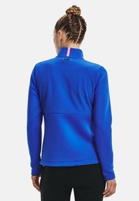 Kvinna med flätat hår som bär en blå långärmad träningsjacka, står med ryggen mot kameran mot en enfärgad bakgrund.