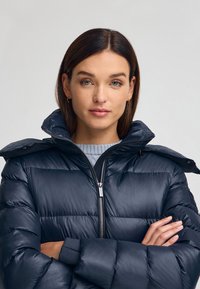 Granatowa kurtka puchowa z wysokim kołnierzem, zamek błyskawiczny i elastyczne mankiety. Posiada pikowane sekcje oraz gładką, błyszczącą fakturę.