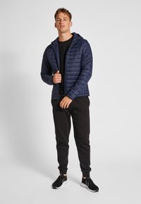 Giacca puffer blu navy con cappuccio, design trapuntato, cerniera frontale, indossata sopra una maglietta nera e pantaloni della tuta neri, abbinata a scarpe da ginnastica nere.