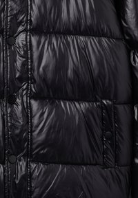 Giacca a piumino nera con una texture lucida e trapuntata. Presenta una chiusura frontale con bottoni e una tasca a toppa, mostrando un design compatto e moderno.