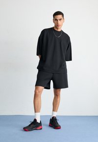 Černé oversized tričko s krátkým rukávem a odpovídající šortky, s hladkou texturou. Černé tenisky s červenými akcenty, bílé ponožky do kotníků.