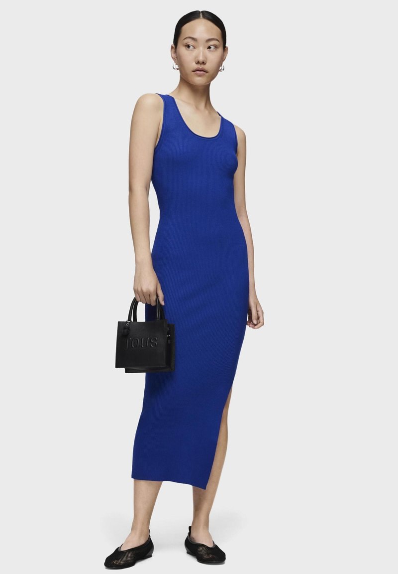 Niebieska sukienka midi z prążkowanym wzorem, dekoltem w kształcie łódki i bocznym rozcięciem, zestawiona z małą czarną torebką o strukturze i czarnymi płaskimi butami z siateczki.