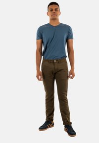 Camisa de algodón azul con escote en V, pantalones ajustados marrones y zapatillas azules con acentos naranjas. Modelo de pie recto frente a un fondo liso.