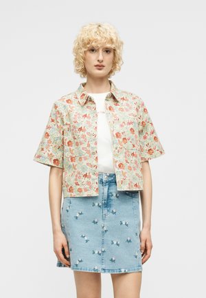 Ung person med kort krøllet blondt hår iført en blomsterprintet kortærmet skjorte over en hvid top og et lyseblåt denimskørt med små blomsterbroderier.