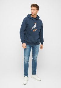 Marineblauwe hoodie van katoen, met een afbeelding van een vogel op een skateboard. Gecombineerd met lichtblauwe jeans en witte sneakers.