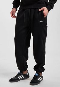Sorte joggebukser laget av myk stoff, med snor i livet, sidelommer og teksturerte detaljer langs sidene.