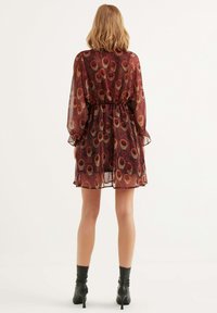 Durchsichtiges rotes Kleid mit einem gemusterten Design in Gold und Schwarz, elastischem Taillenbund, langen Ärmeln und gerafften Bündchen. Getragen mit schwarzen High Heels Stiefeln.