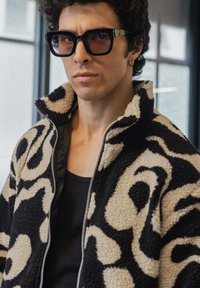 Veste en polaire avec un motif abstrait noir et beige, dotée d'une fermeture éclair sur le devant et d'un col haut. Des lunettes de soleil à monture foncée rehaussent le look.