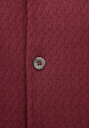 Bordeauxroter Strickstoff mit strukturiertem Muster und einem einzelnen runden, durchsichtigen grauen Knopf mit vier Löchern, der nahe der Mitte angenäht ist.