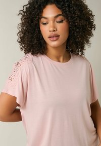 Ljust rosa kortärmad t-shirt med rundad halsringning och intrikata spetsdetaljer på axlarna, tillverkad av mjukt, elastiskt tyg.