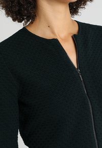 Ciemnozielona kurtka o fakturze z zapięciem na zamek, z okrągłym dekoltem i subtelnymi geometrycznymi wzorami na materiale.