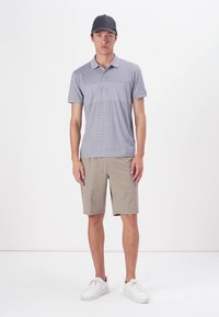 Graues kariertes Poloshirt mit kurzen Ärmeln, kombiniert mit beigen Shorts und weißen Sneakern. Eine schwarze Mütze rundet das legere Outfit ab.
