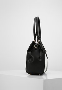 Musta rakenne-tasku, jossa on kaksinkertaiset kantokahvat, valkoiset ja harmaat tekstuuripinnat, lepää valkoisella jalustalla yksinkertaista taustaa vasten.