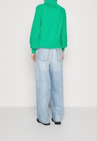 Pull tricoté vert avec un col montant et des manches texturées, associé à un jean denim bleu clair à jambes larges. Tissu texturé et coupe décontractée.