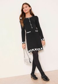 Rochie neagră cu mâneci lungi, cu garnitură decorativă albă, nasturi în față și tiv evazat. Asortată cu colanți negri și pantofi loafer din lac.