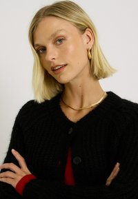 Cardigan noir en maille avec de gros boutons, décolleté large et texture côtelée. Porté par-dessus un haut rouge, avec un collier en or en accent.