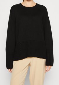 Svart stickad tröja med ribbad textur, breda ärmar och lös passform, i kombination med beige byxor. En enkel och mångsidig design.