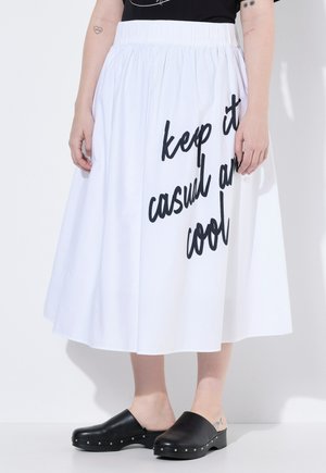 Person trägt einen weißen Rock mit schwarzer Schrift „keep it casual and cool“, schwarzes Oberteil, schwarze Slip-On-Schuhe mit Nieten und sichtbare Arm-Tattoos.