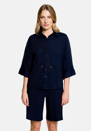 Femme aux cheveux mi-longs portant une chemise bleu marine boutonnée avec une taille à cordon de serrage et un short assorti jusqu'aux genoux, debout sur fond blanc.