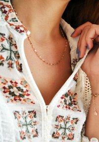 Delicada cadena de oro con un diseño retorcido, descansando sobre un escote adornado con bordados florales en rojo, verde y crema sobre una camisa blanca.