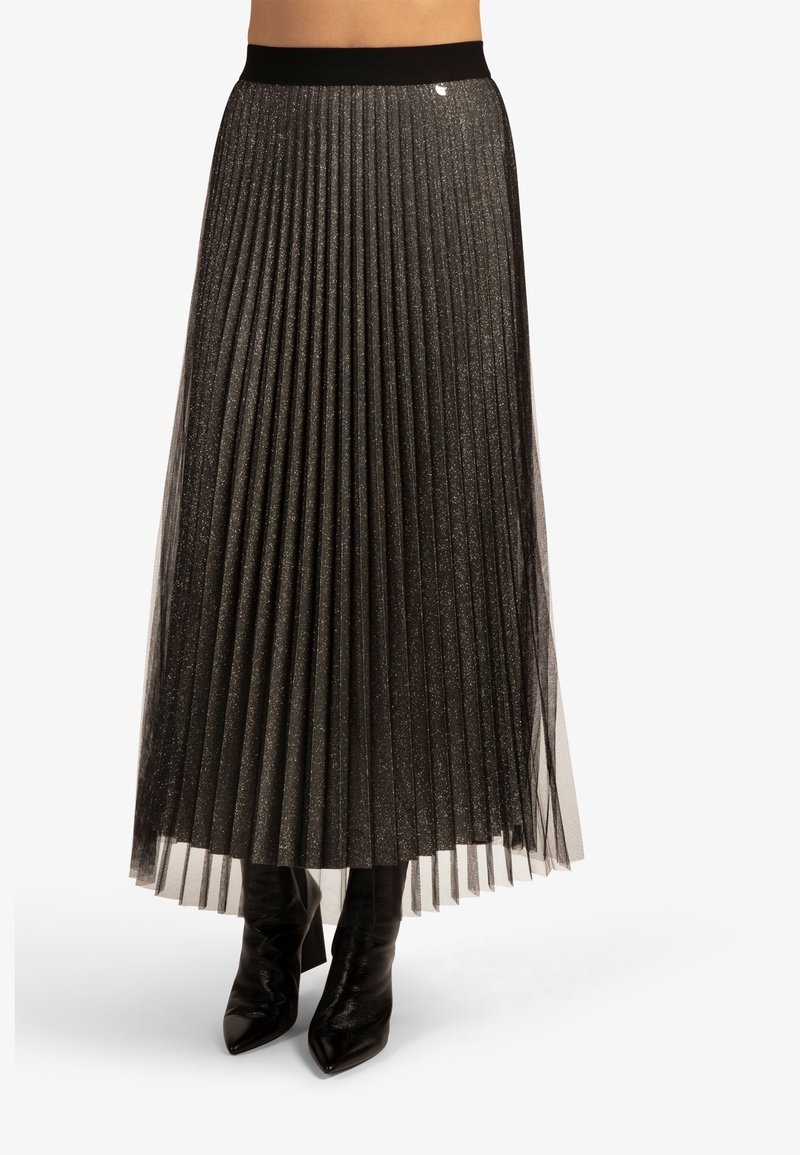 Jupe plissée noire en tissu transparent à texture scintillante, dotée d'une ceinture noire et associée à des bottes noires montantes jusqu'aux genoux.