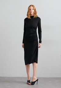 Robe noire à manches longues avec des détails froncés, longueur genoux, tissu doux, coupe ajustée, encolure arrondie, assortie à des talons ouverts noirs.