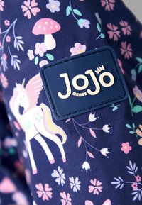 Tessuto blu scuro con motivi floreali rosa e viola, un unicorno bianco con ali rosa e una toppa blu navy con la scritta "JoJo maman bébé."