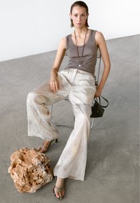 Lichtbruine tanktop, losse crèmekleurige broek met een glans, zilveren sandalen met banden, zittend op een metalen stoel naast een gestructureerde steen. Zwarte tas.