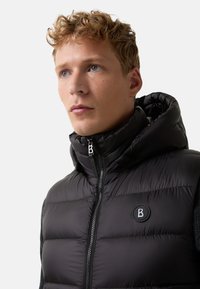 Czarna kamizelka puchowa z wysokim kołnierzem, z zamkiem błyskawicznym i naszywką z logo. Materiał wydaje się gładki, z pikowaną teksturą.