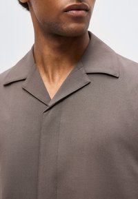 Άνδρας με μπεζ πουκάμισο με λαιμόκοψη με εγκοπή, εμφανιζόμενος από τον λαιμό έως το πάνω μέρος του στήθους, με κλειστά χείλη, κοιτάζοντας ελαφρώς πλάγια.