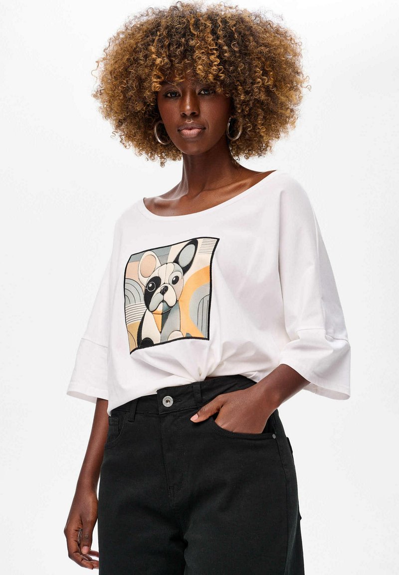Biała, oversize t-shirt z graficznym nadrukiem psa w kolorach szarości, czerni i pomarańczy. Posiada szerokie rękawy i miękką teksturę.