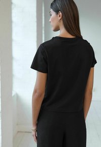 Haut noir à manches courtes avec un col rond, présentant une texture lisse et un design court. La vue arrière montre des lignes épurées et un minimum de détails.