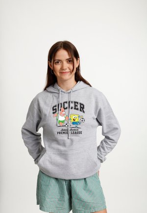 Młoda kobieta w szarej bluzie z kapturem z nadrukiem SpongeBoba i Patricka grających w piłkę nożną oraz zielonych szortach w paski, stojąca na jednolitym białym tle.