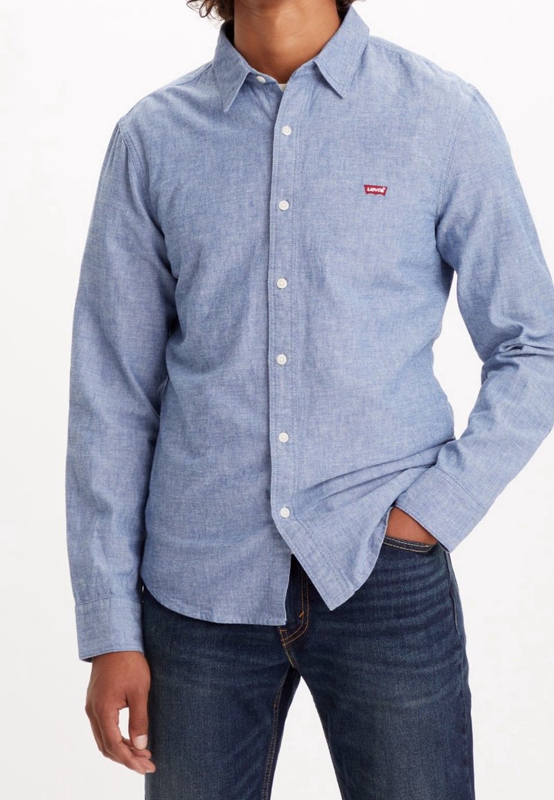 Camicia button-up azzurro chiaro in tessuto chambray con colletto classico, maniche lunghe e un piccolo logo rosso sul petto sinistro. Abbinata a jeans scuri.