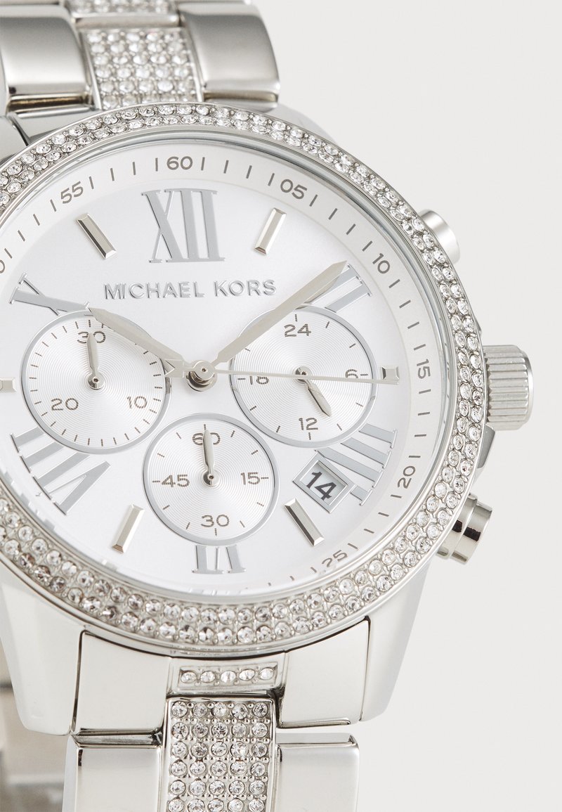 Montre analogique en argent avec un boîtier rond, un cadran blanc, des index en chiffres romains, trois sous-cadrans, et une lunette et un bracelet ornés de cristaux.