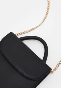 Musta tekonahkainen käsilaukku, jossa on yläkahva ja kultaketjuolkainten. Ominaisuuksiin kuuluu läppä suljenta ja sileä pinta. Kompakti ja suorakulmainen muoto.