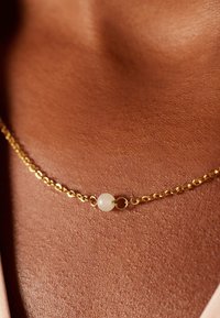 Gouden ketting met een ovaal schakeldesign, met een witte ronde kraal in het midden, die tegen een textuurover huid ligt.