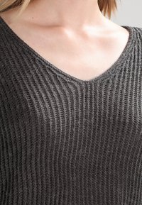 Pull gris en tricot avec un col en V, présentant une texture côtelée et un tissu doux et souple qui s'ajuste étroitement autour du cou.